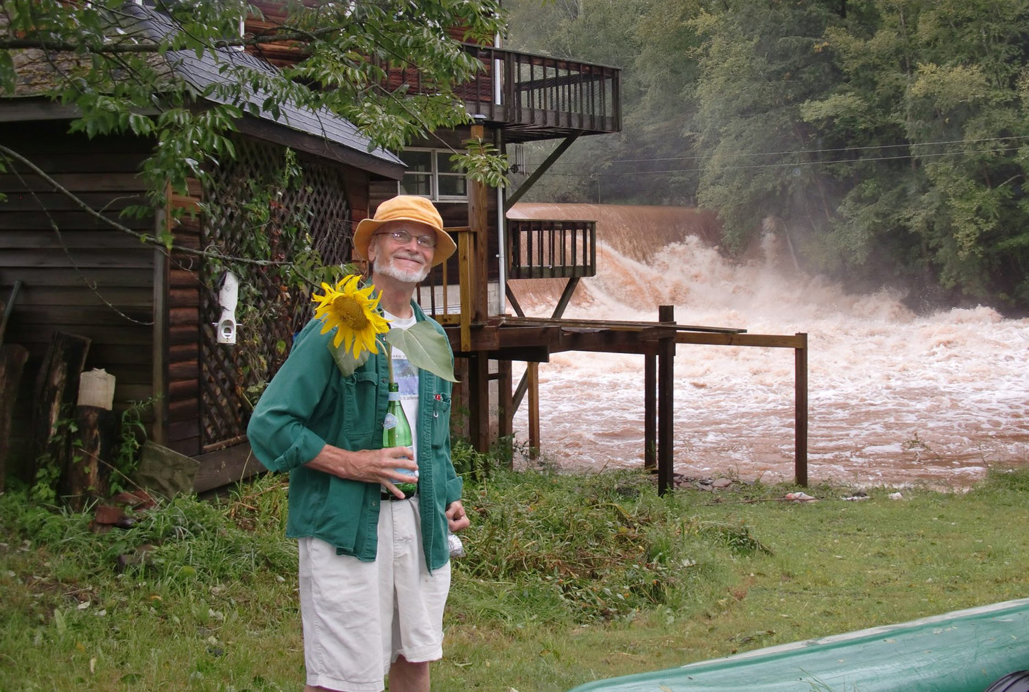 Malcolm Brown, holding a bright yellow sunflower, stands before the entrance to Jeffersonville Hydroelectric in 2011. In the background is the spillway of the Lake Jefferson dam, the source of power for the company’s renewable energy generators. ..
